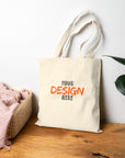 Beige tote bag with 'Your Design Here' text on a wooden surface with a plant in the background
