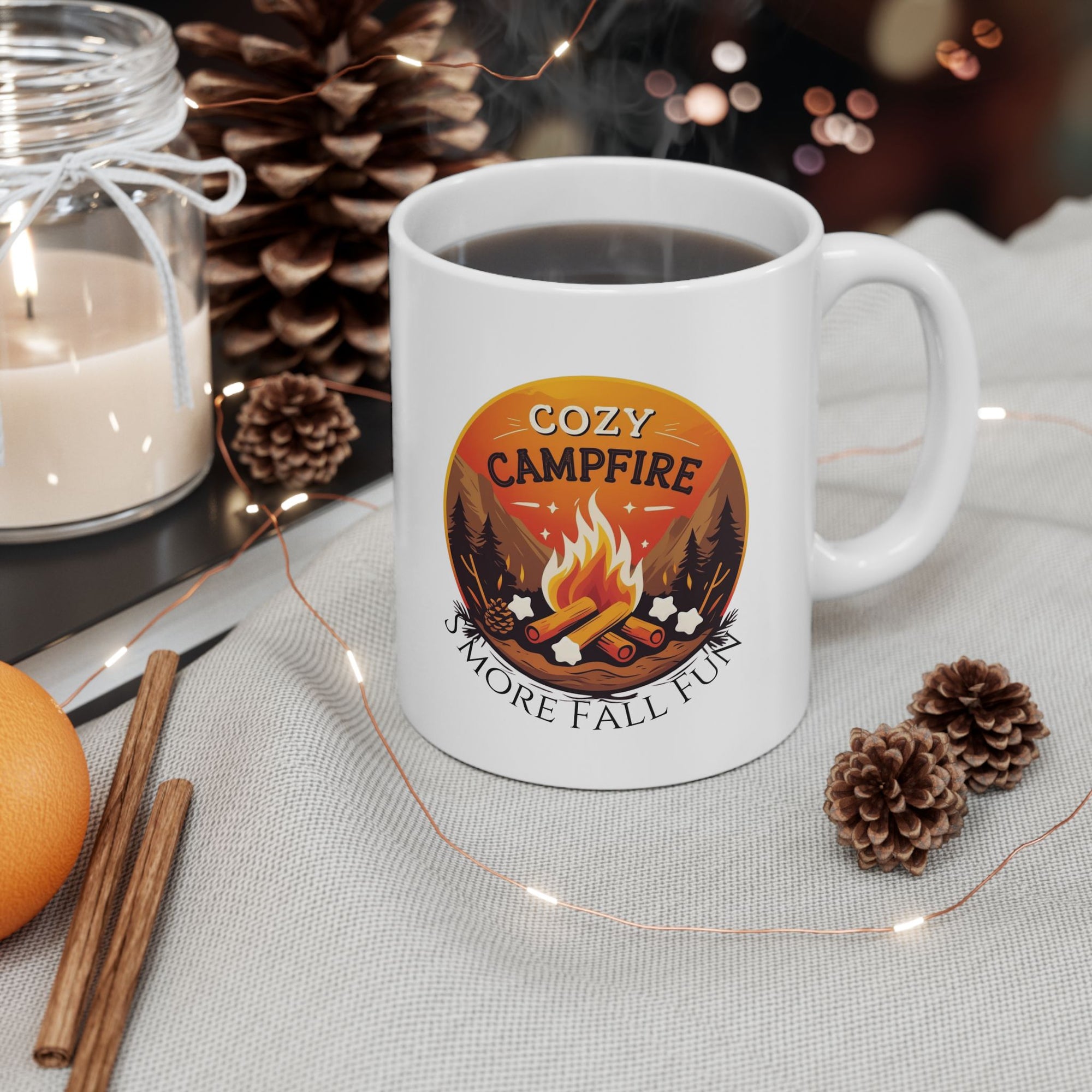 White mug with 'Cozy Campfire' design on a table with candles, oranges, and pinecones.