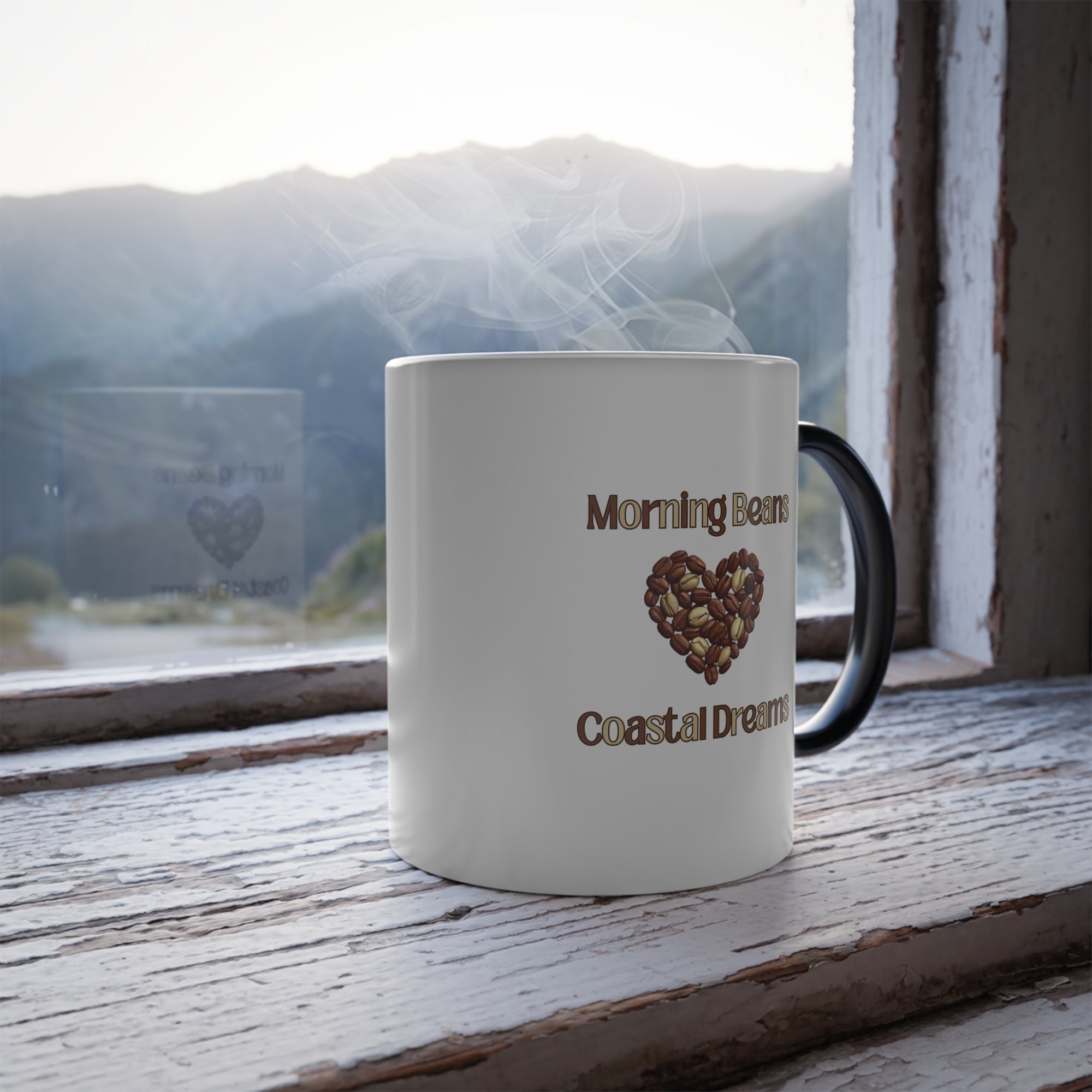 White mug with 'Morning Beans Coastal Dreams' text and heart design on a wooden surface with a window view of mountains.