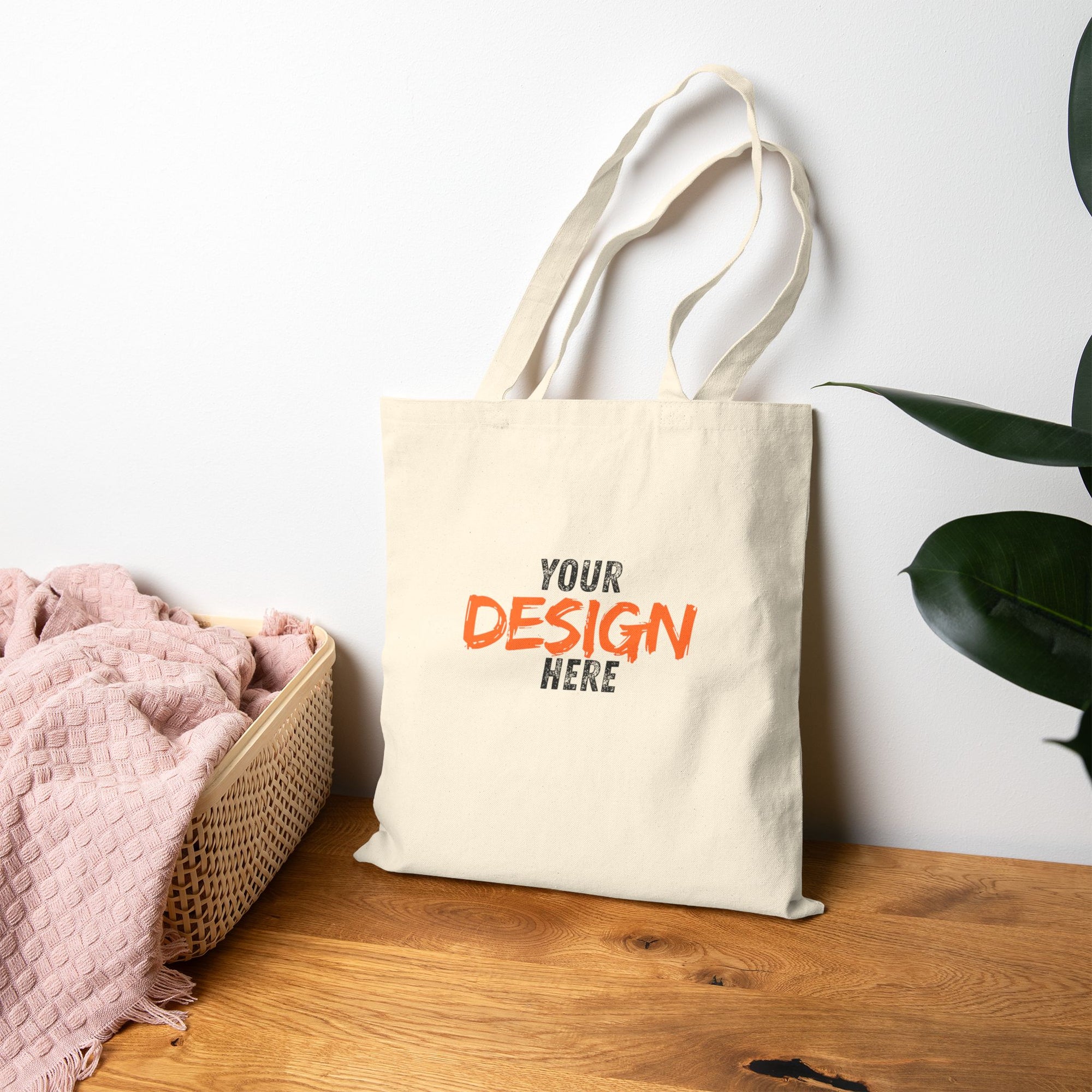 Beige tote bag with 'Your Design Here' text on a wooden surface with a plant in the background