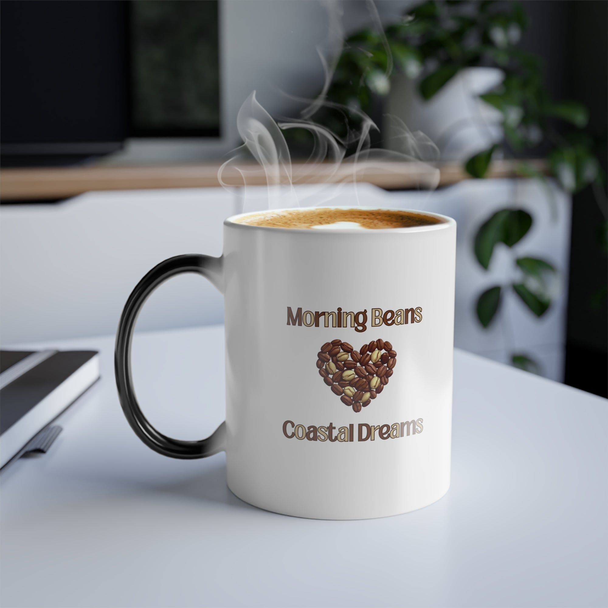 White mug with black handle and 'Morning Beans Coastal Dreams' text and heart design on a desk.